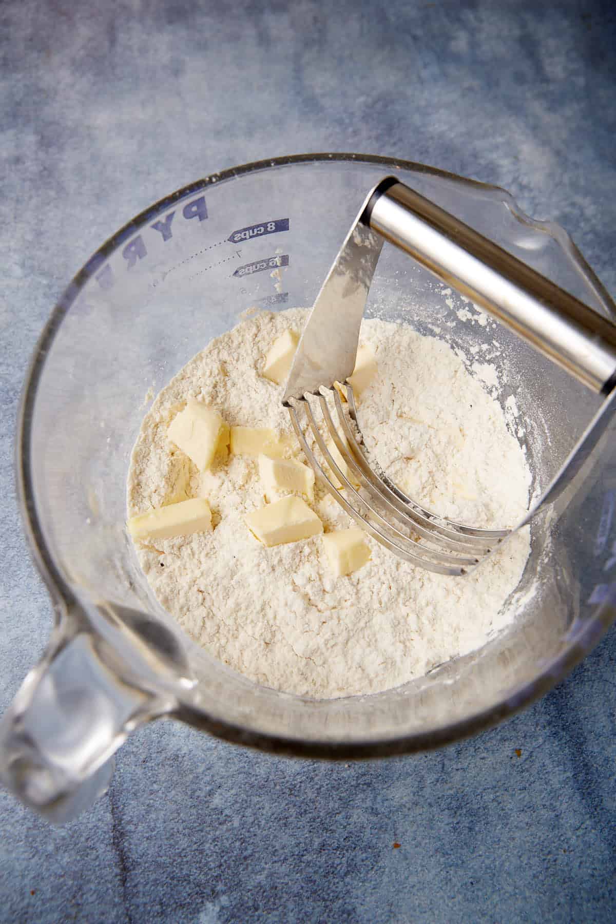 A glass mixing bowl containing flour and small chunks of butter. A metal pastry cutter is placed inside the bowl, with some of the butter and flour mixture clinging to it. The bowl is set on a blue, textured surface.