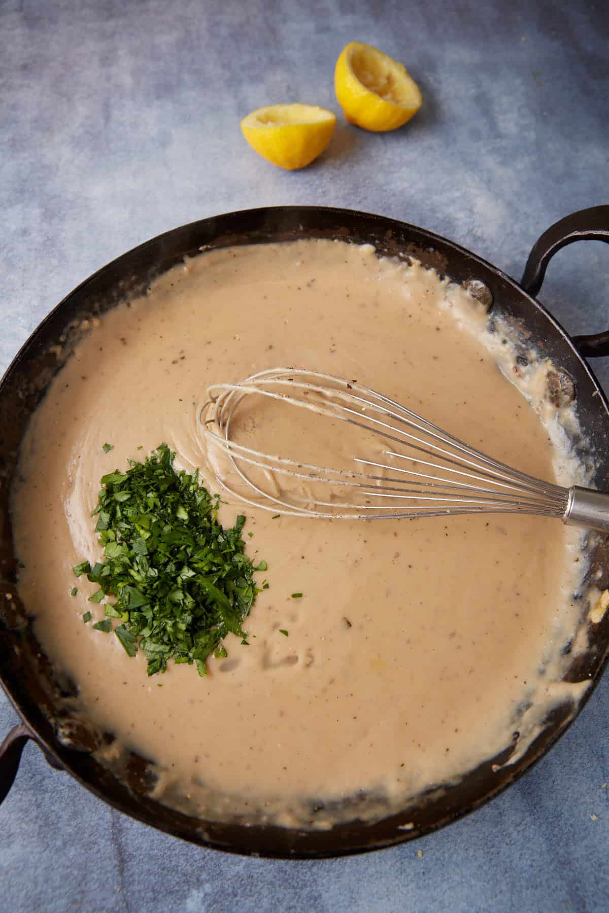 A skillet filled with creamy white sauce being whisked, topped with freshly chopped parsley. Two lemon halves are visible in the background on a blue-gray surface.