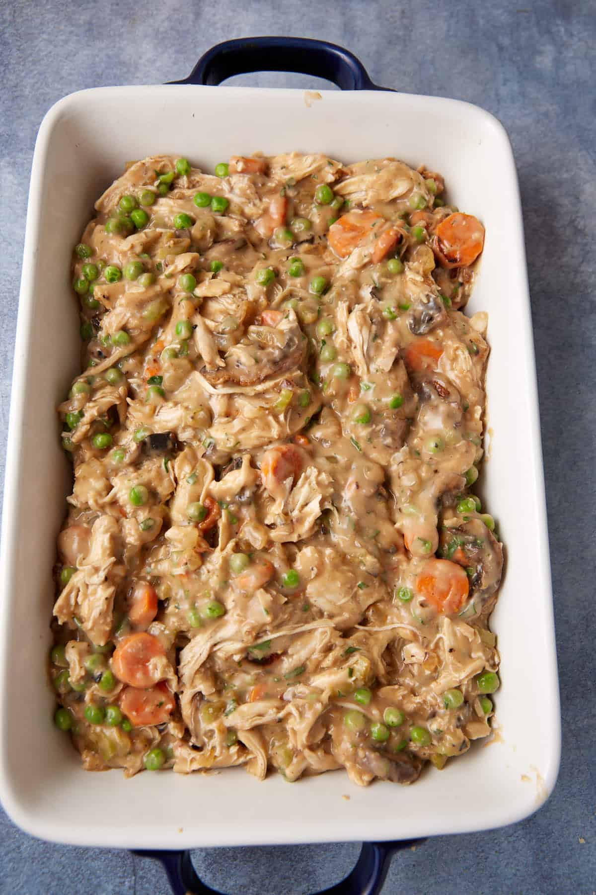 A white rectangular baking dish filled with a creamy chicken mixture, featuring shredded chicken, peas, sliced carrots, and mushrooms in a thick, savory sauce. The dish has handles on either side and rests on a grey surface.