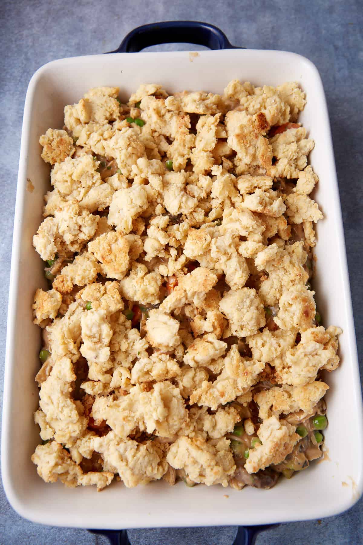 A baking dish filled with golden-brown, crumbly biscuit topping over a mixture of peas, carrots, and a creamy sauce, resembling a pot pie or casserole, placed on a blue-gray surface.