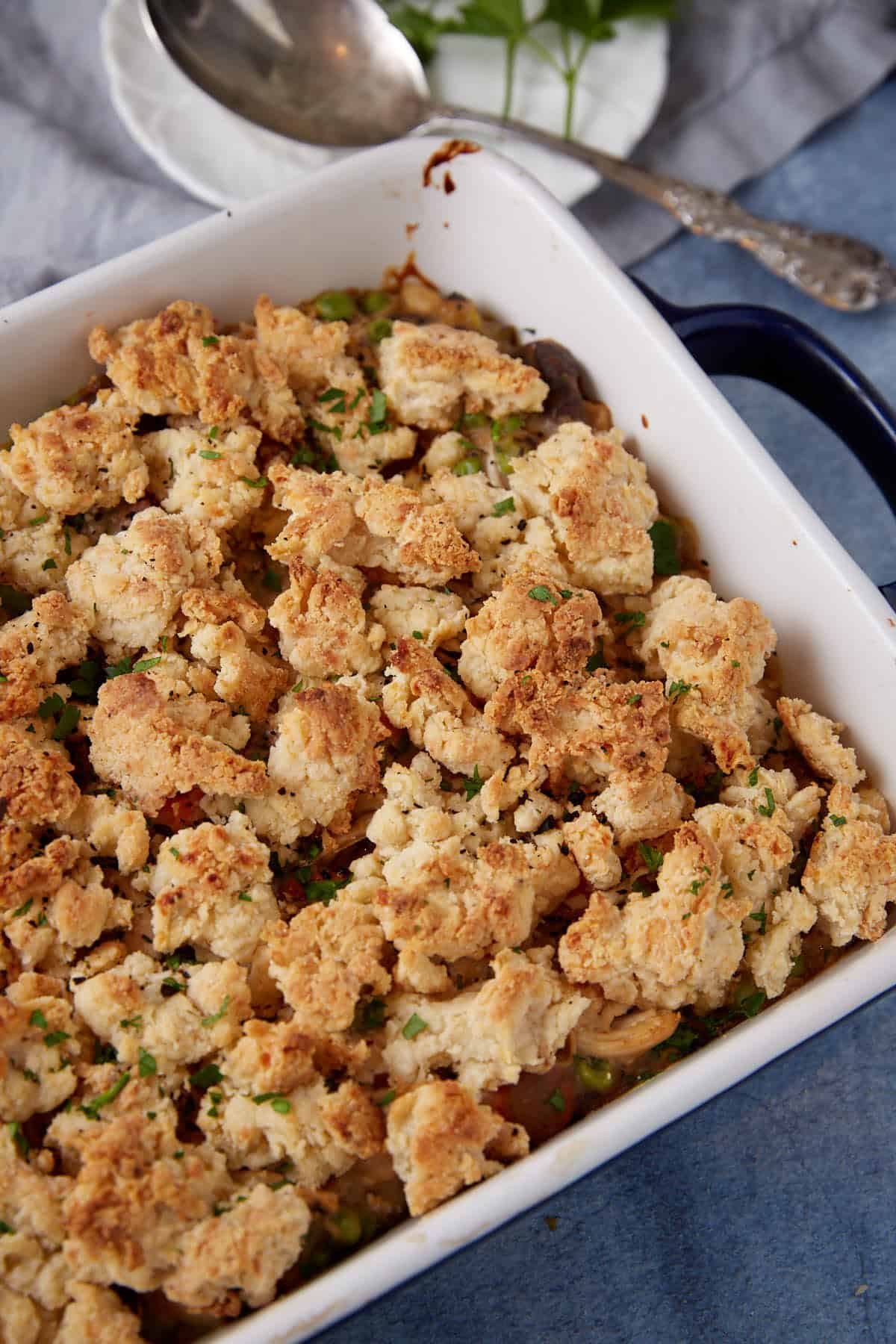 A baked casserole dish filled with a crumbly, golden brown topping and scattered herbs. The dish appears to contain a mixture of vegetables and possibly meat underneath the topping. In the background, there is a spoon and a partial view of some green garnishes on a small plate.