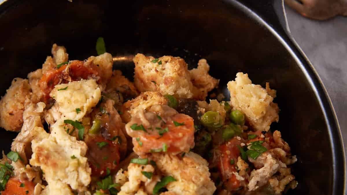 A black bowl filled with chunks of golden-brown stuffing mixed with pieces of vegetables and herbs. A casserole dish and a fork lie in the background on a gray tablecloth, adding a cozy, rustic feel to the scene.