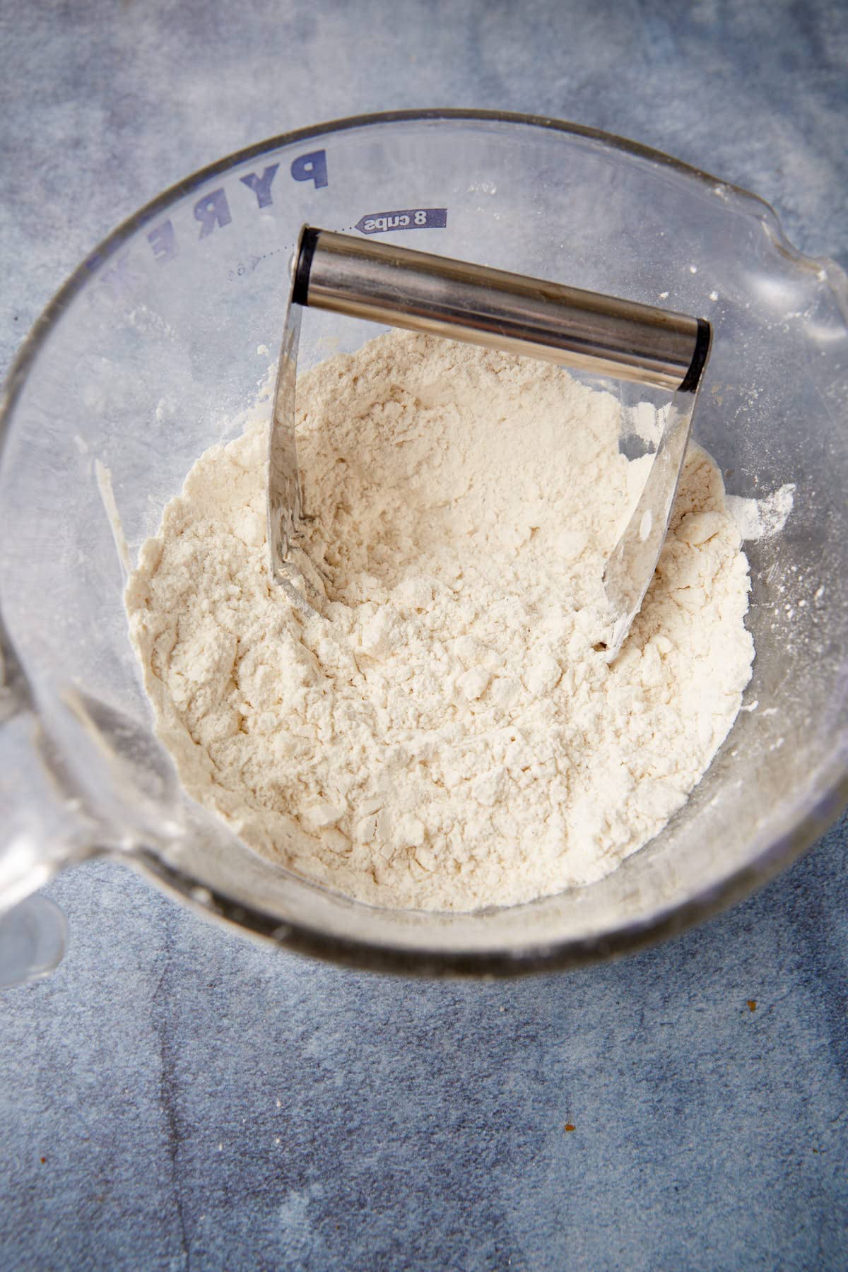 A clear Pyrex bowl filled with flour mixture is on a blue surface. A metal pastry cutter is partially submerged in the flour, ready to blend the ingredients further.