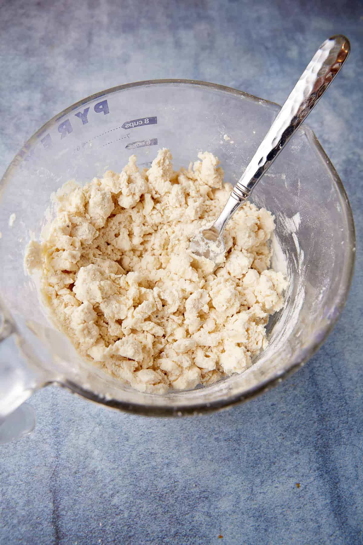 A glass Pyrex measuring bowl containing a mixture of kneaded dough with a metallic fork placed inside. The background is a blue and white textured surface. The dough appears to be in the initial stages of mixing, characterized by a crumbly texture.