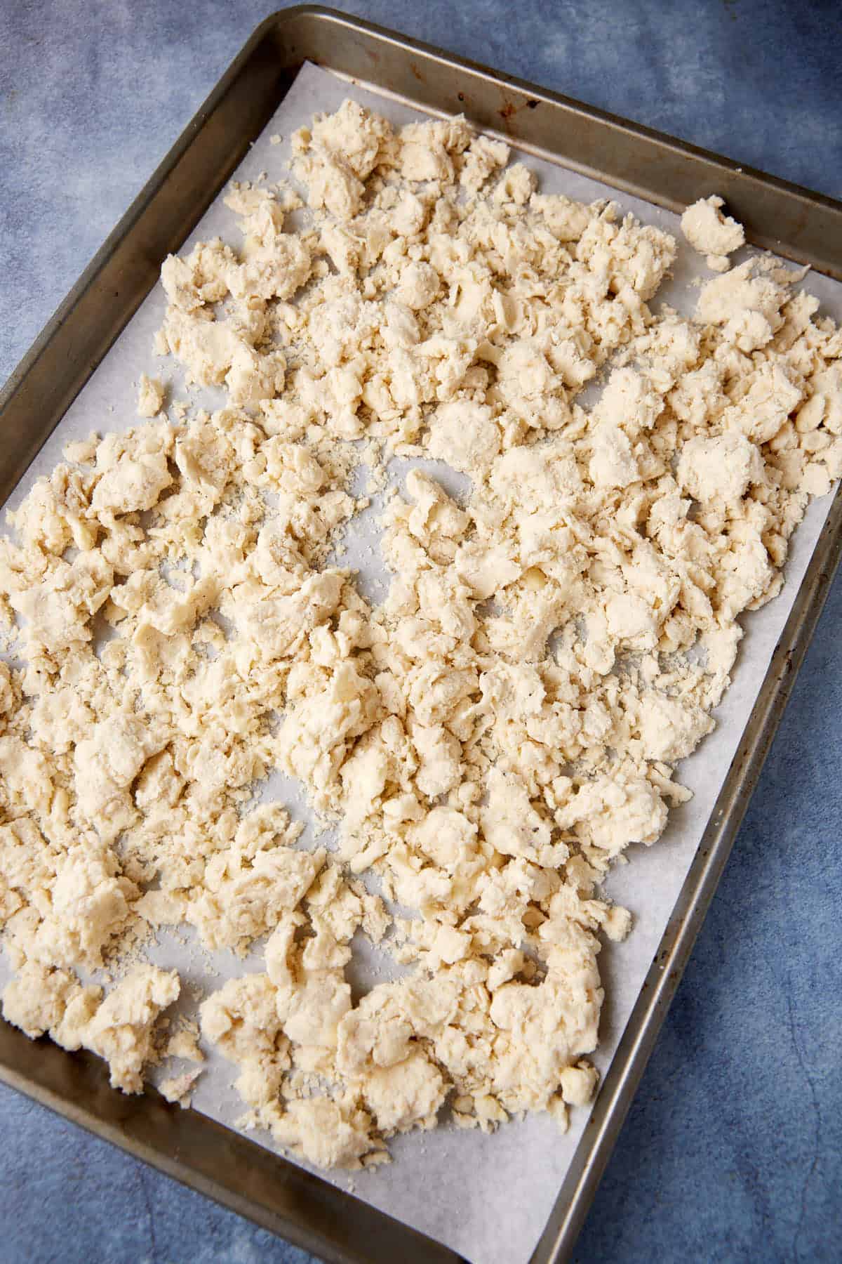 A large, rectangular baking sheet with a layer of crumbled dough spread unevenly over a sheet of parchment paper. The background surface is a textured blue-gray color.