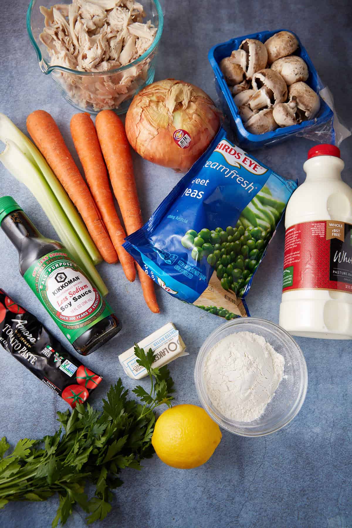 A collection of cooking ingredients spread on a blue surface, including cooked shredded chicken, carrots, celery, an onion, mushrooms, frozen peas, whole milk, soy sauce, flour, butter, tomato paste, fresh parsley, and a lemon.