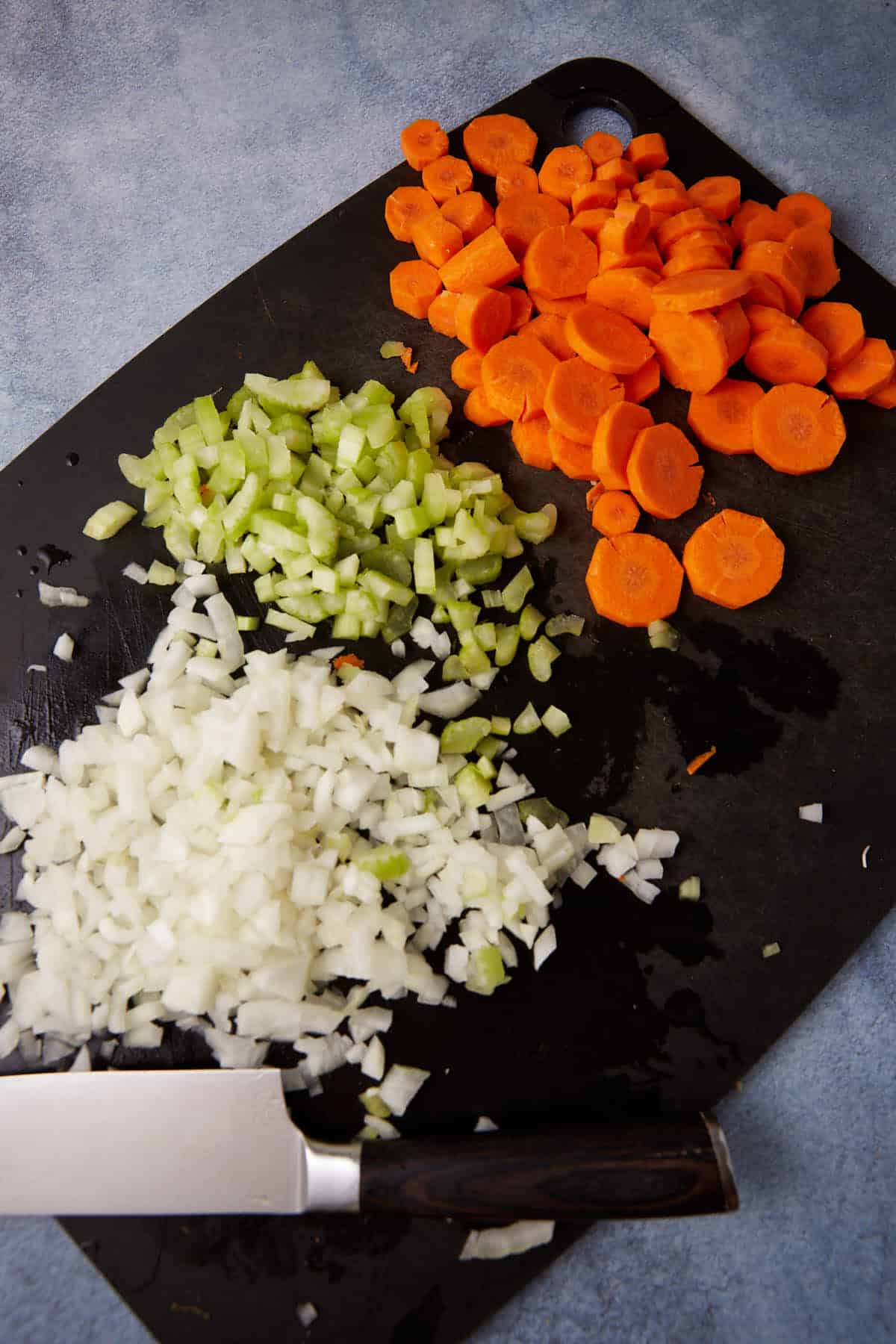 A black cutting board displays chopped carrots, celery, and onions, each in a separate pile. A chef's knife with a black handle lies next to the vegetables. The background is a lightly textured blue surface.