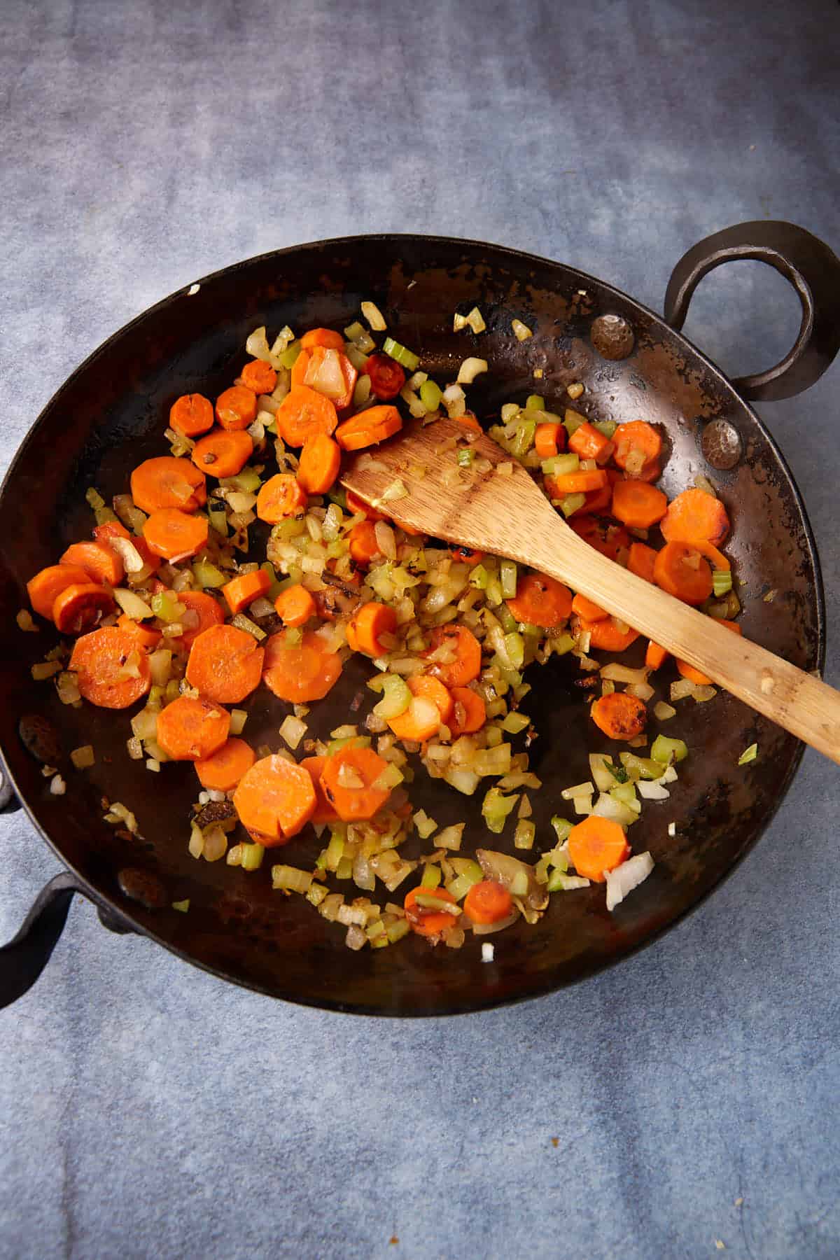 A frying pan with sautéed vegetables, including chopped carrots, celery, and onions, being stirred with a wooden spatula. The pan rests on a gray surface.
