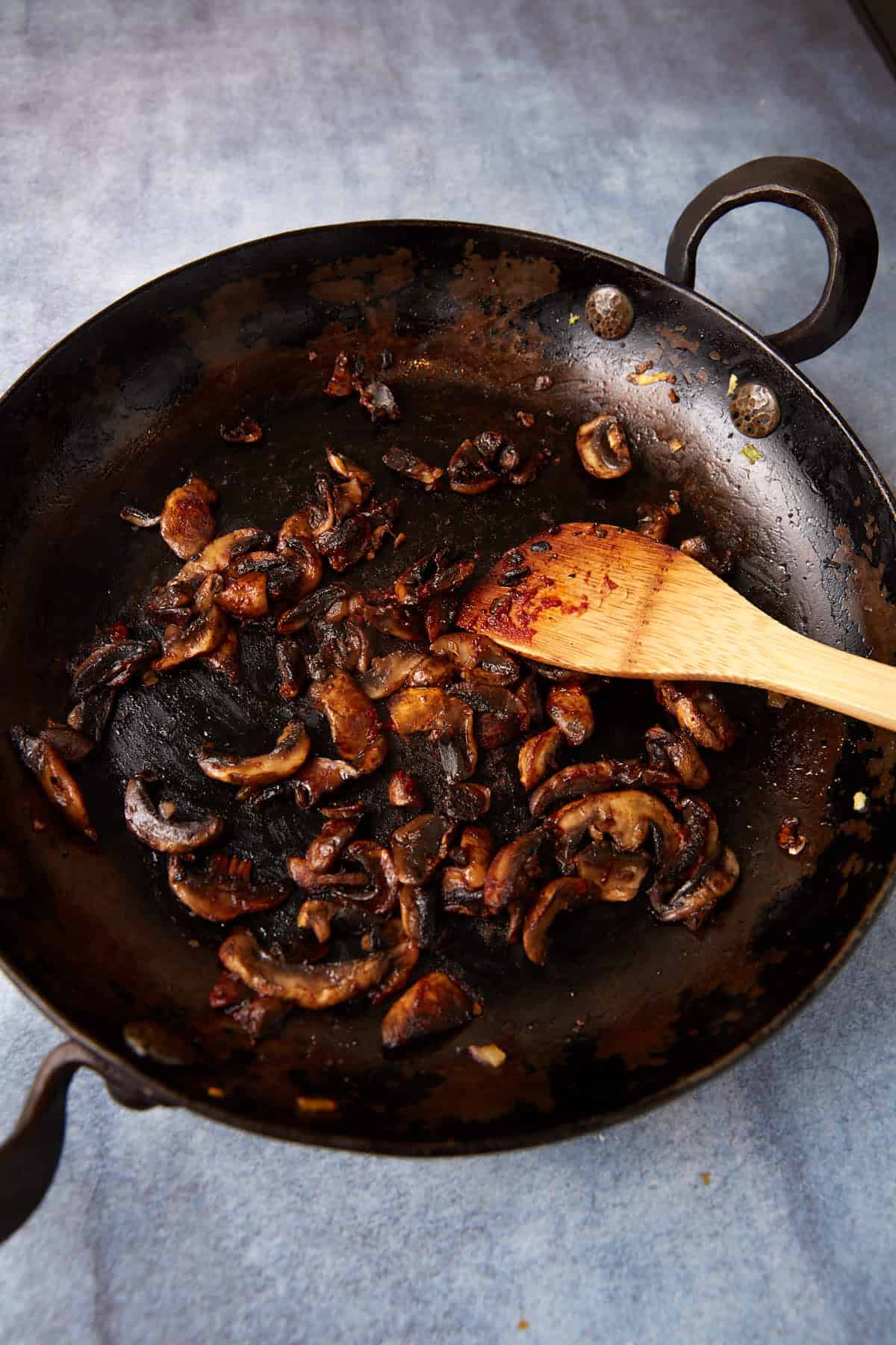 A black cast iron skillet filled with sautéed mushrooms sits on a stovetop. The mushrooms are browned and have a caramelized appearance. A wooden spatula rests in the skillet, indicating they are being stirred or cooked.