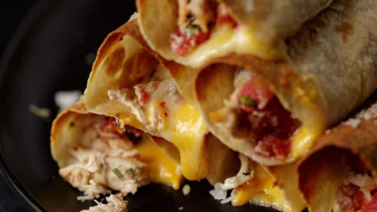 Close-up of crispy rolled tortillas filled with shredded chicken, melted cheese, and diced tomatoes, stacked on a dark plate—a delicious addition to your favorite chicken breast recipes.