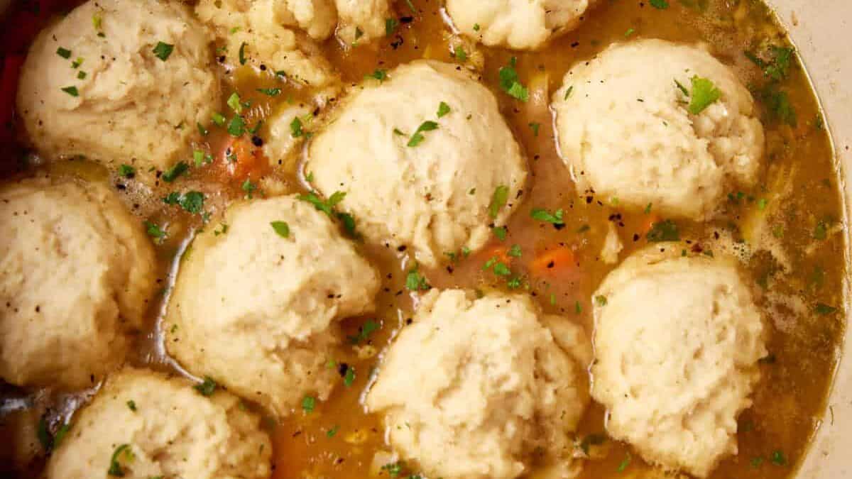 A pot filled with chicken and dumplings. The dish features round, golden dumplings resting in a savory broth with visible carrots, herbs, and bits of chicken. The surface is garnished with chopped parsley. The pot sits on a blue-gray countertop.