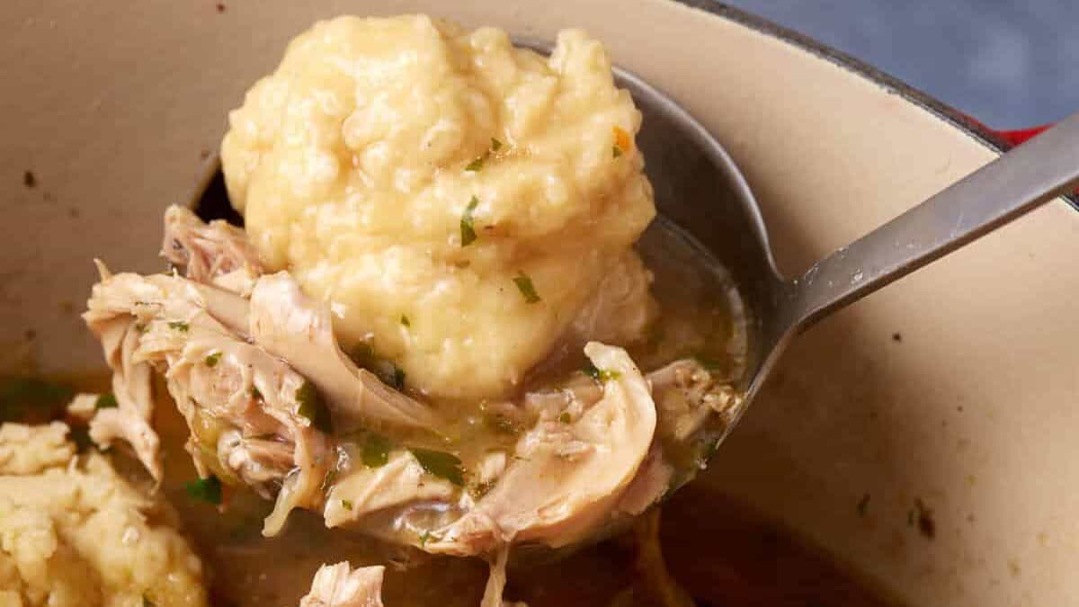A pot of chicken and dumplings with chunks of chicken, carrots, and herbs in a flavorful broth. A ladle is lifting a dumpling and some chicken out of the pot, showcasing the hearty comfort food.