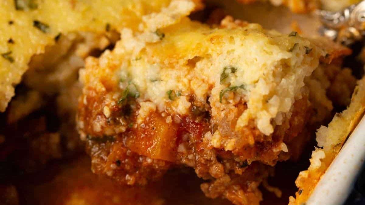 Close-up of a baked dish in a casserole, featuring a golden, herb-speckled topping. A serving has been scooped out, revealing layers of ground meat in a chili poblano tomato-based sauce underneath. The dish looks hearty and freshly made.