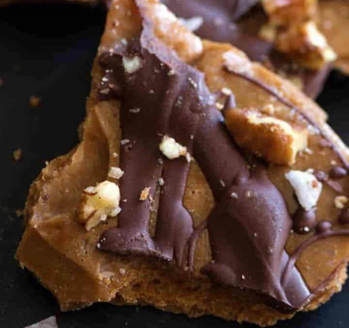Close-up of chocolate toffee pieces scattered on a dark surface. The toffee is topped with a layer of hardened chocolate and sprinkled with chopped nuts. Some pieces are broken into irregular shapes, and the overall appearance is appetizing and crunchy.
