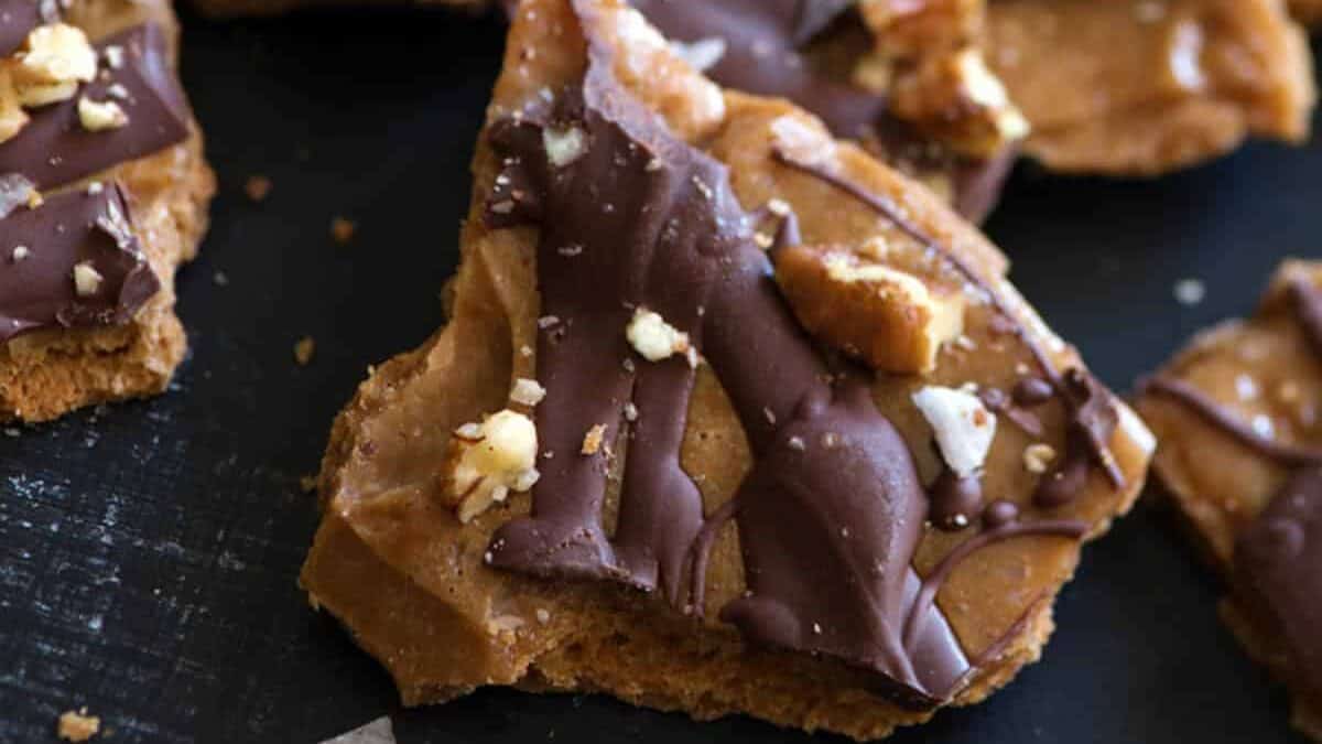 Close-up of chocolate toffee pieces scattered on a dark surface. The toffee is topped with a layer of hardened chocolate and sprinkled with chopped nuts. Some pieces are broken into irregular shapes, and the overall appearance is appetizing and crunchy.