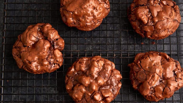 A batch of rich, chocolatey brownies in the form of cookies is displayed on a black wire cooling rack. The cookies are evenly spaced in a grid pattern, showcasing their cracked, glossy tops and dense, fudgy interior.