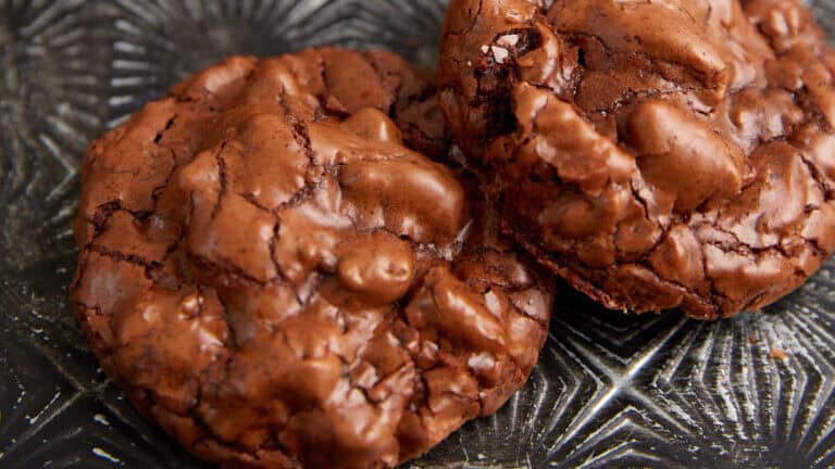 Two rich, chocolate cookies with a crinkled surface displayed on a textured metallic tray. The cookies have a fudgy appearance with chunks of chocolate embedded in them. Blurred in the background, there is a glass of milk and another cookie.