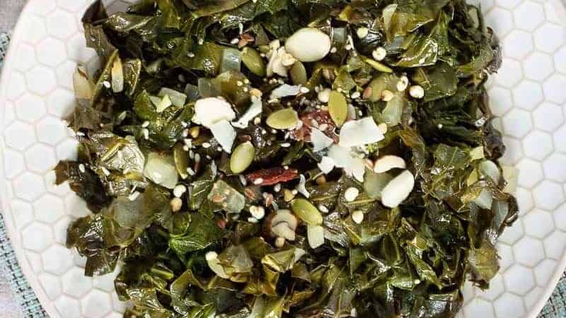 A plate of cooked greens, garnished with various seeds and nuts, is presented on a white honeycomb-patterned plate. Two silver forks are placed on the plate. The plate rests on a textured light blue and white fabric.