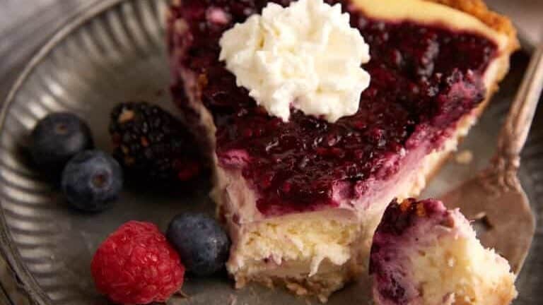 A close-up of a slice of cheesecake topped with berry compote and a dollop of whipped cream on a metal plate. A fork holds a piece of the cake, and fresh blueberries and raspberries are on the side. A gray cloth and a bowl of raspberries are in the background.