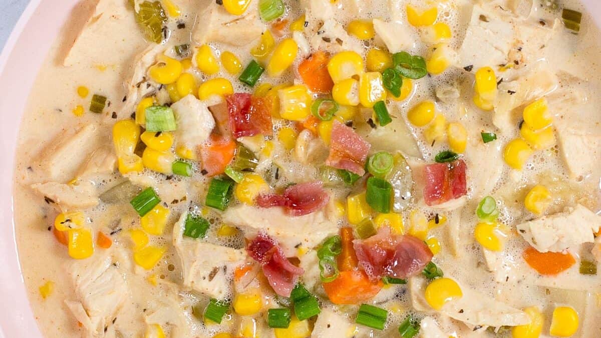 A creamy chicken and corn chowder garnished with green onions and pieces of bacon is served in a light-colored bowl. The soup is filled with chicken chunks, corn kernels, diced carrots, and potatoes. Additional bowls of bacon bits and chopped green onions are nearby.