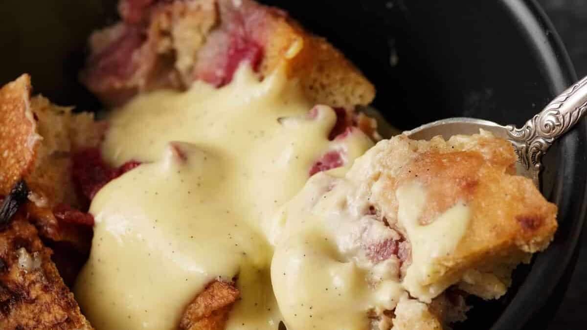 A close-up view of a bread pudding topped with a generous amount of creamy vanilla sauce. The pudding consists of bread pieces and visible chunks of red fruit, likely berries, served in a black bowl with a spoon scooping a portion.