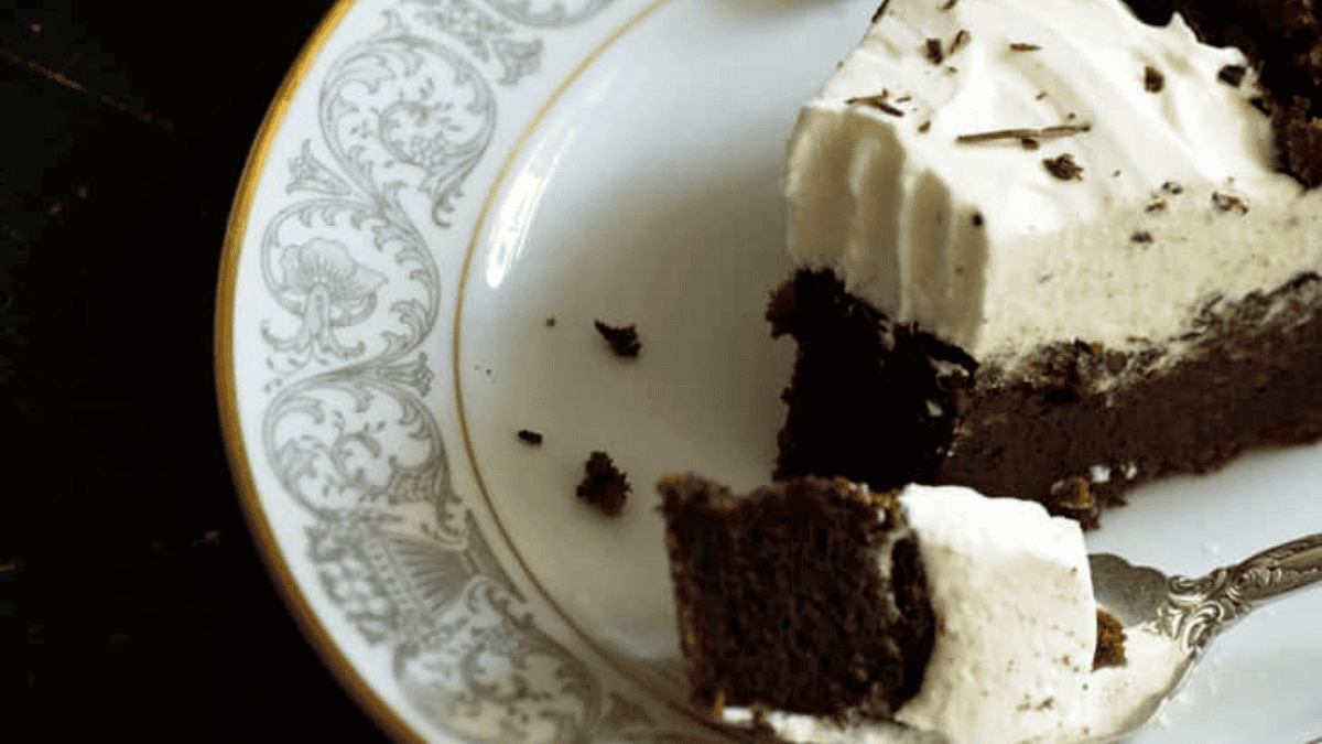 A slice of chocolate cake with creamy white frosting on a decorative white and silver plate. The rest of the cake is in the background on a dark surface, with a piece cut out and garnished with chocolate shavings. Crumbs are scattered around.
