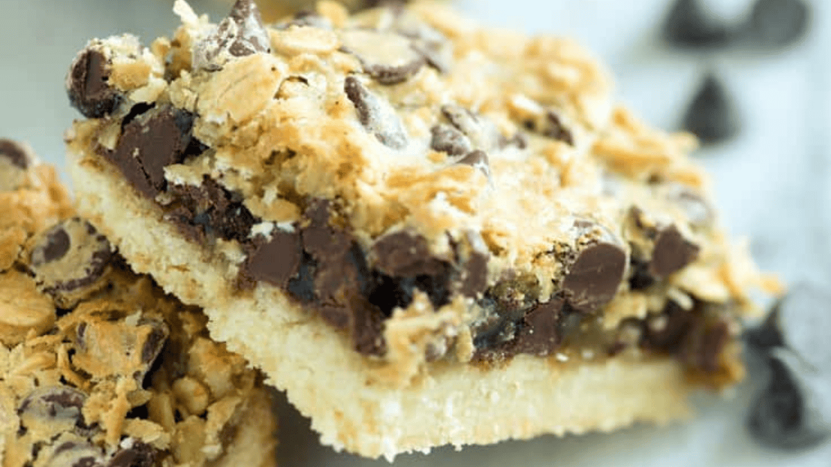 A close-up of a dessert bar with layers of chocolate chips, sweetened condensed milk, crumbled topping, and a crust base. The bars are stacked in the background, with a few chocolate chips scattered on a marble surface. The bars have a crumbly texture and appear freshly baked.
