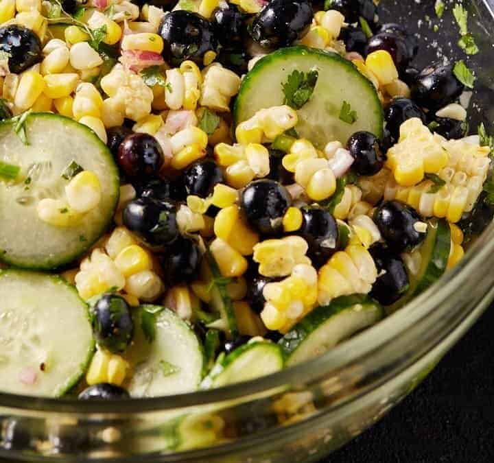 A clear glass bowl of corn salad with blueberries.