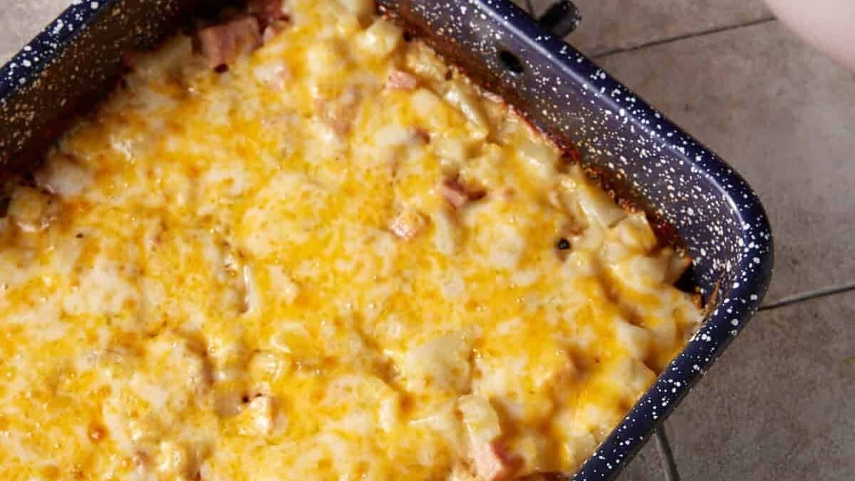 A rectangular blue baking dish filled with a cheesy baked potato casserole sits on a tiled countertop. To the upper left, there is a stack of white plates with silver forks resting on top. In the upper right, there is a bowl of mixed fruit, including grapes and strawberries.
