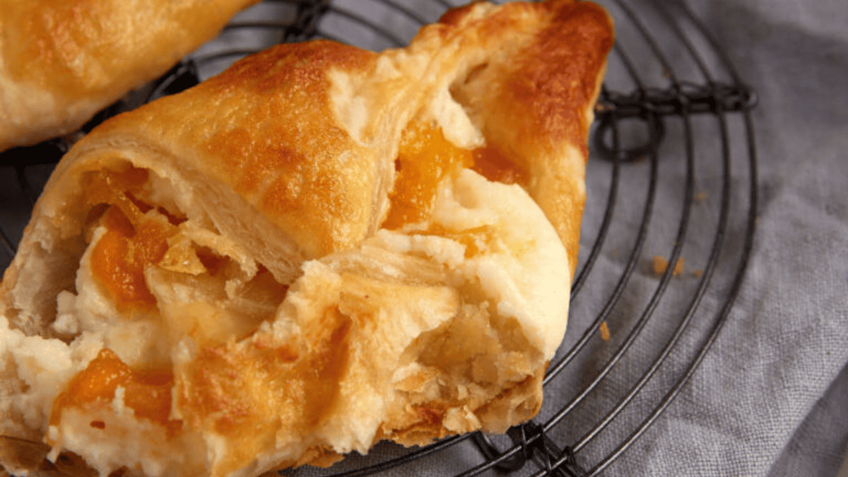 A close-up of a flaky pastry with visible apricot filling on a cooling rack placed on a grey cloth. In the back, there is a small bowl with more apricot pieces. Some crumbs and a pastry fragment are in the foreground.