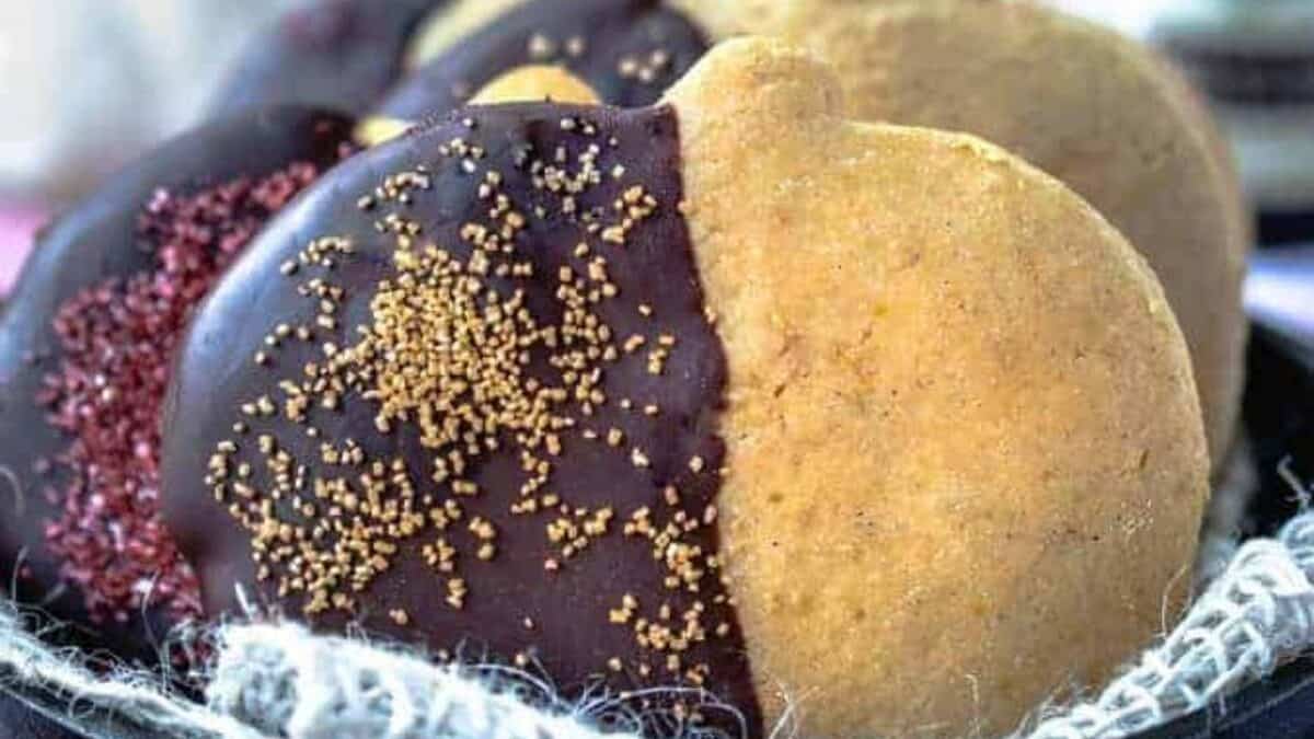 Close-up of round cookies partially dipped in dark chocolate and decorated with colorful sprinkles. The cookies are placed in a black dish lined with a white net. The background is blurred with hints of purple and white.