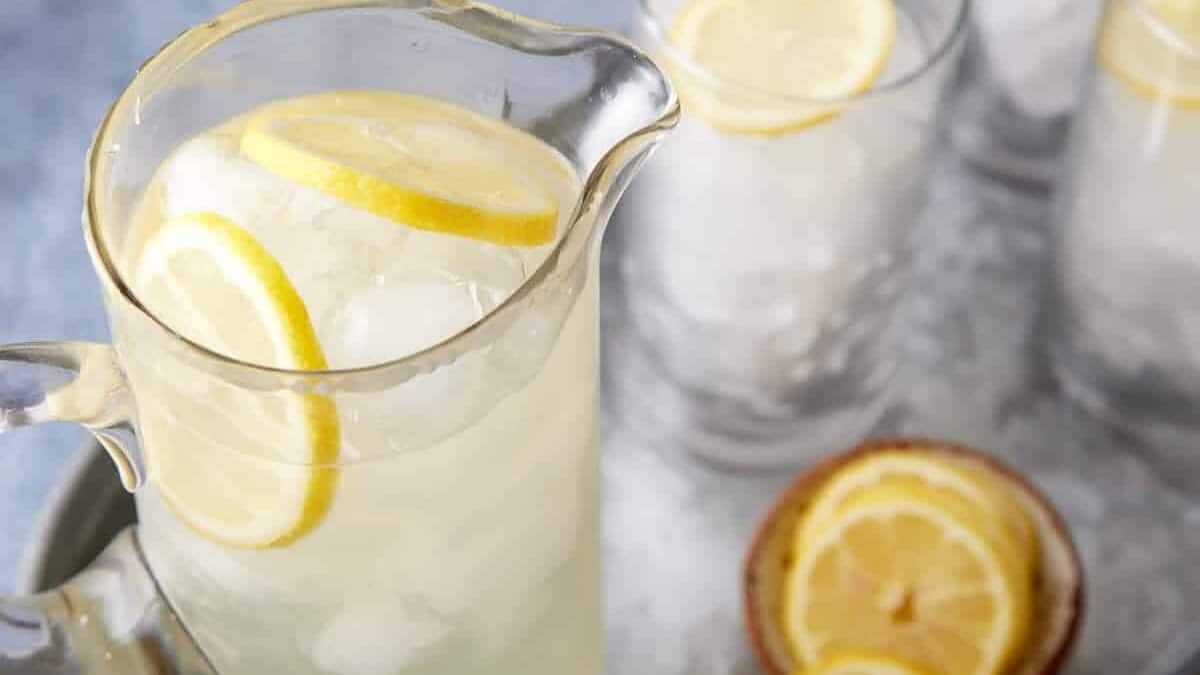 A pitcher filled with lemonade and lemon slices sits on a tray alongside three empty glasses with ice and lemon slices. Two halved lemons are also on the tray, and the background features a light blue surface.