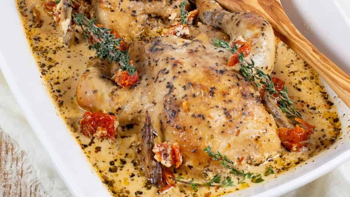 A white rectangular dish contains cooked chicken breasts covered in a creamy herb sauce. The dish, showcasing one of our cherished family recipes, is garnished with sun-dried tomatoes and fresh thyme sprigs. A wooden spoon rests on the side, and the dish is set on a rustic wooden surface with a cloth underneath.