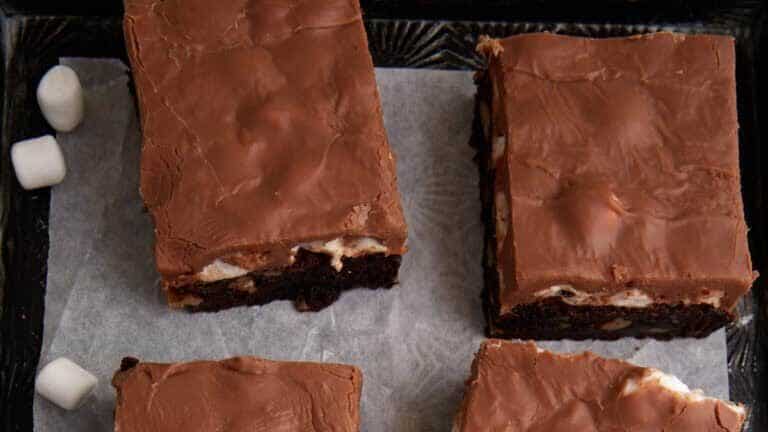 An image of six chocolate-topped marshmallow brownies arranged on a sheet of parchment paper. The brownies are rectangular and have a smooth, shiny chocolate layer on top with visible marshmallow layers beneath. Small marshmallows are scattered around them.