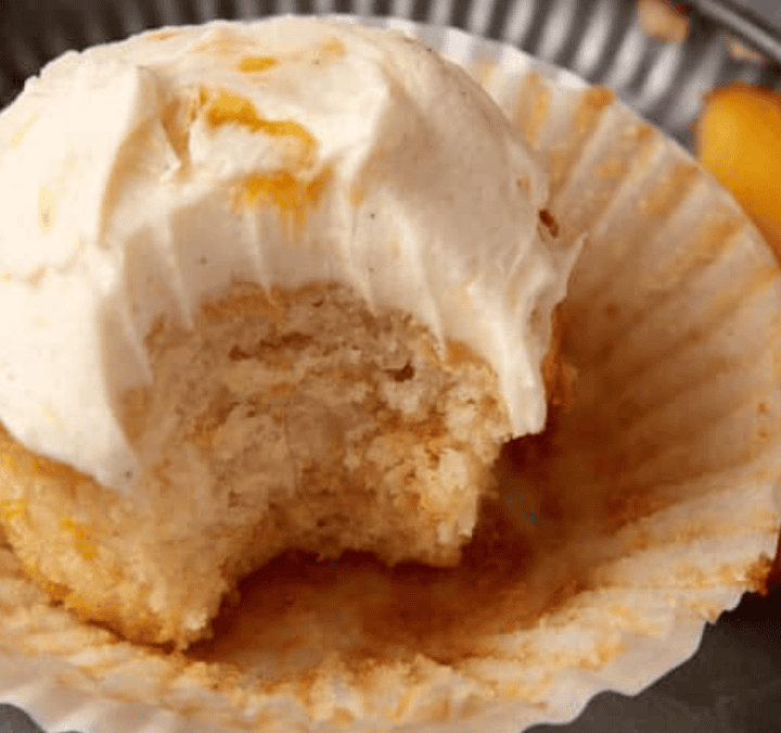 A vanilla cupcake with creamy white frosting sits in a partially peeled paper liner, showing a bite taken out. Two peach slices are on the plate—perfect inspiration for Easy Dessert Recipes.