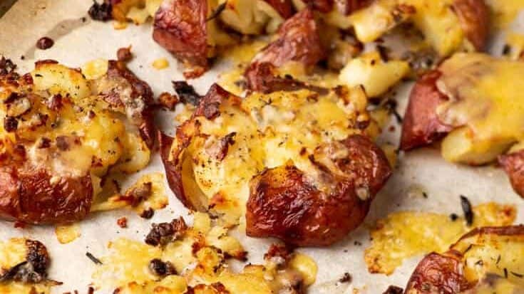 A baking sheet covered with parchment paper holds several baked smashed red potatoes. Topped with melted cheese and sprinkled with crispy bits, possibly bacon or breadcrumbs, they showcase a golden-brown, appetizing appearance perfect for your collection of potato recipes.