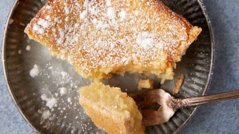 A slice of powdered sugar-dusted cake is on a grey ceramic plate with a silver fork. Nearby, a partially sliced cake rests in a baking dish, and a gold-rimmed dish containing additional powdered sugar sits on a cooling rack with a cloth. The setup is on a blue surface.
