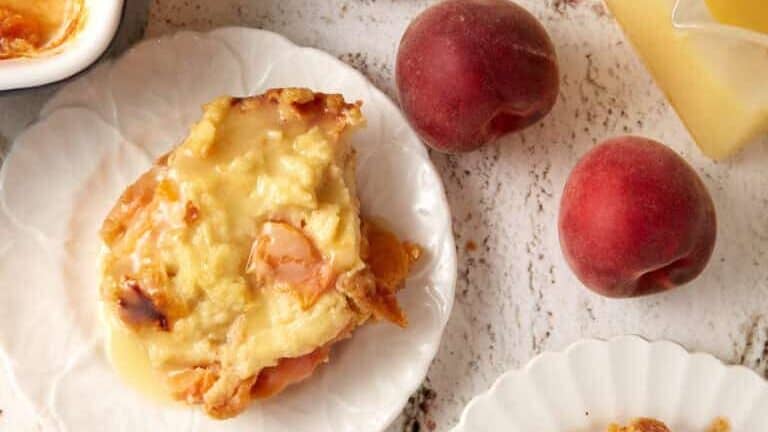 A top-down view of peach cobbler served on three white plates placed on a light-colored, marbled surface. The cobbler appears golden and gooey, accompanied by two whole peaches and a small jug of peach sauce. A baking dish of cobbler is visible in the top left corner.