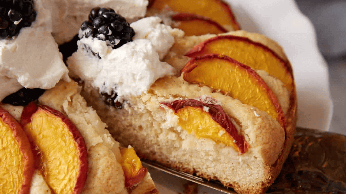 A close-up of a slice of cake topped with sliced peaches, whipped cream, and blackberries, being lifted from the rest of the cake—a tempting treat for anyone who loves dessert recipes. The cake appears moist and fluffy with a golden crust.