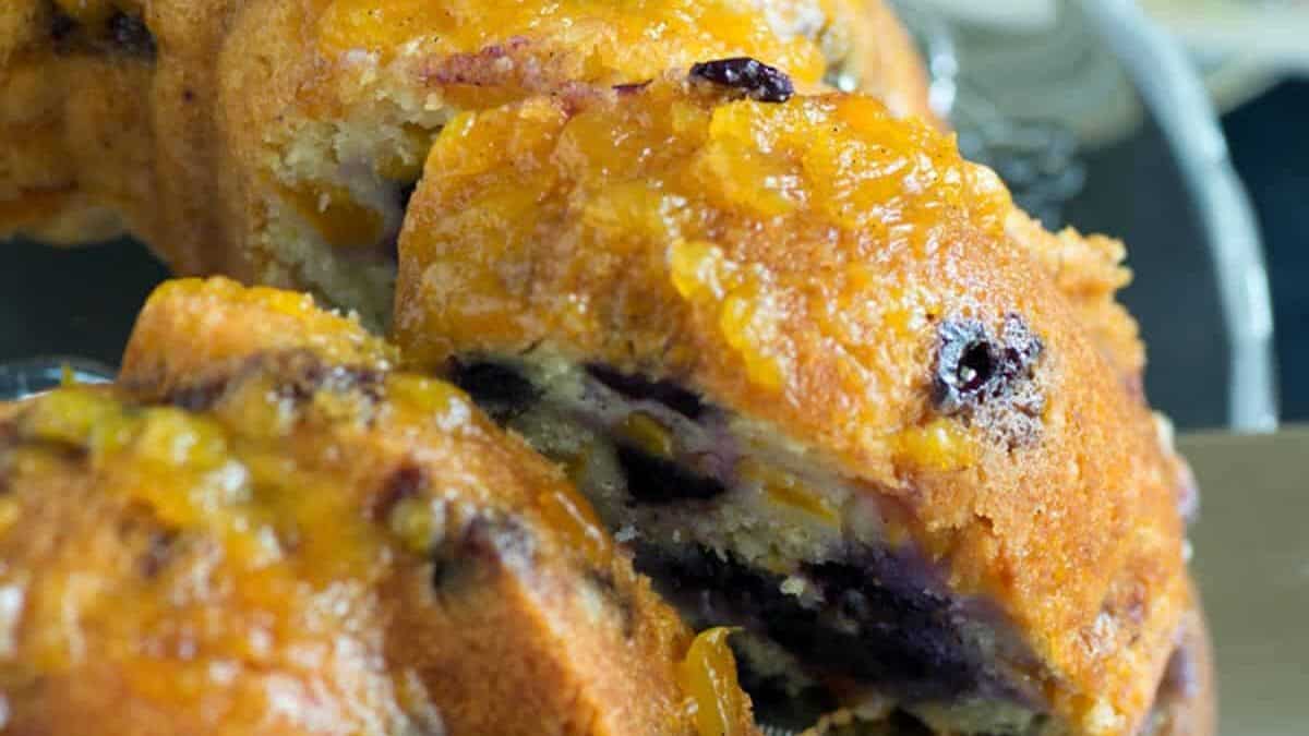 Close-up of a sliced bundt cake on a glass plate, with blueberries and bits of orange zest visible. A stainless steel server is partially inserted under a slice. In the background, a floral napkin and some forks are visible.