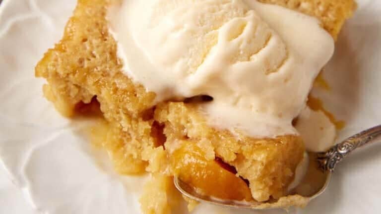 A delectable slice of peach cobbler topped with a generous scoop of creamy vanilla ice cream is served on a white plate. A silver spoon rests beside the dessert. In the background, a whole peach and a glass baking dish with more cobbler—straight from our cherished family recipes—are visible.