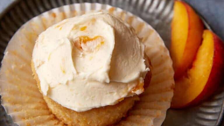 A frosted cupcake with light-colored icing sits on a fluted metal plate, partially unwrapped from its paper liner. Beside it are two peach slices. In the background, more cupcakes rest on a cooling rack. The background surface is light gray.