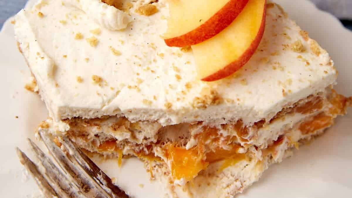 A slice of dessert featuring layers of creamy filling and visible peach slices sits on a white plate. Two peach slices garnish the top, highlighting the allure of cream cheese desserts. A fork rests beside the treat. Another piece is visible in the background, next to a grey napkin and additional forks.