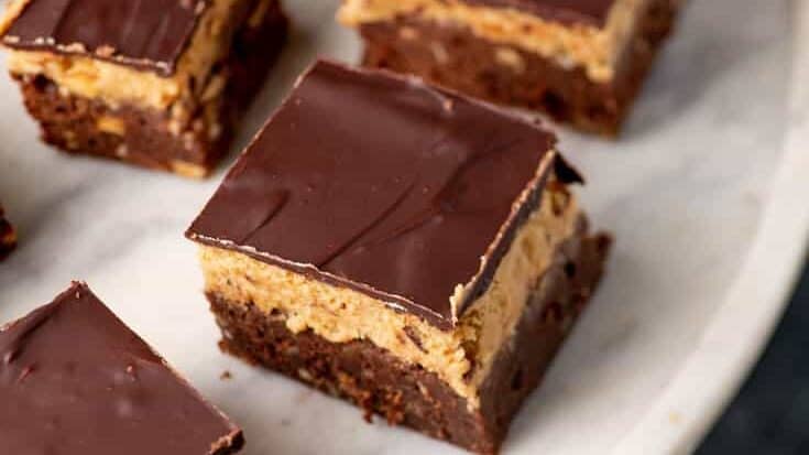 A round white plate holds several neatly cut square desserts. These layered treats feature a chocolate base, a creamy peanut butter middle, and a glossy chocolate top. The layers are distinct, and the chocolate topping appears smooth and rich.