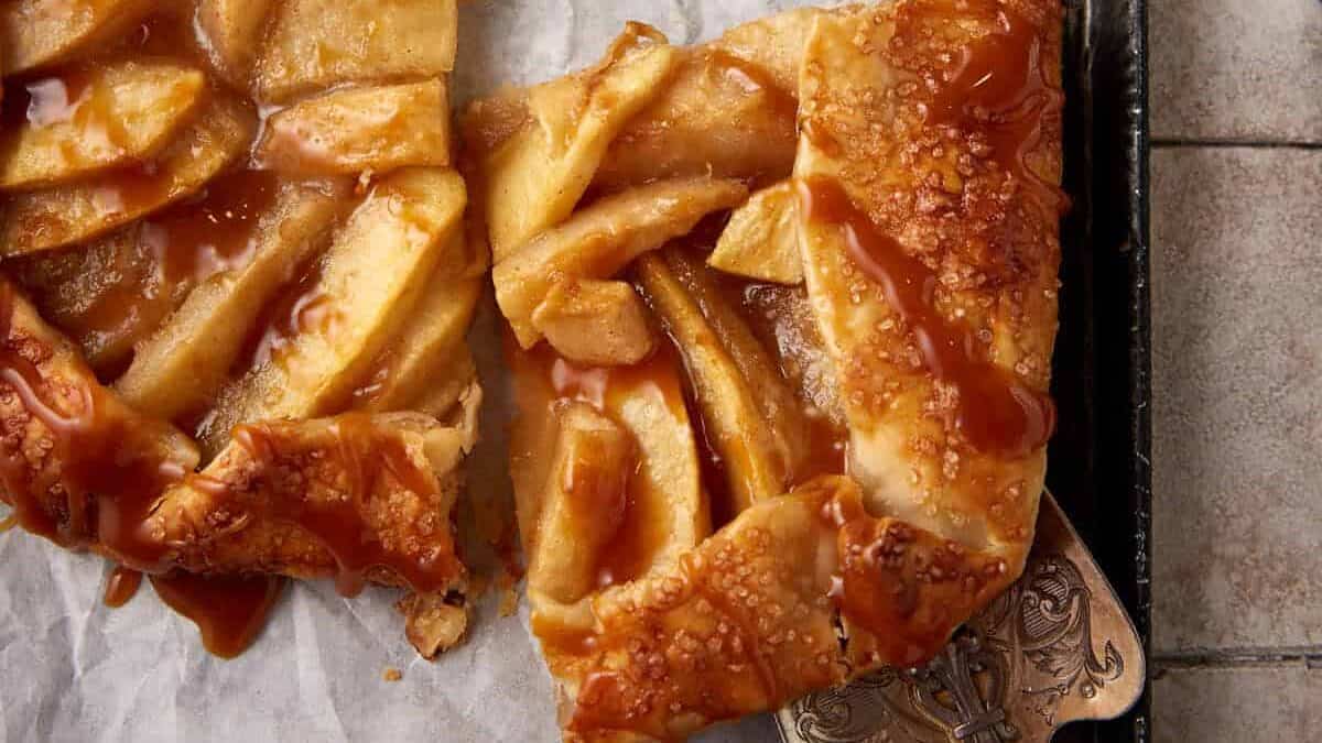 A close-up of a slice of apple pie with a flaky, golden-brown crust and caramelized apples, topped with a shiny caramel drizzle. The pie slice is on a piece of parchment paper with a metal pie server partly visible underneath.