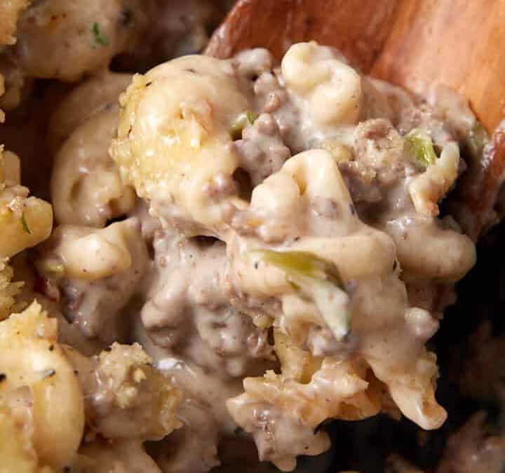 A close-up of a creamy pasta bake with ground meat and a golden breadcrumb topping, reminiscent of a cheesesteak casserole. A wooden spoon is scooping up a portion, revealing the rich, cheesy sauce mixed with pasta and meat underneath the crispy topping, garnished with herbs.
