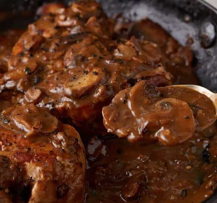 A skillet of pork chops with a spoon full of mushroom gravy.