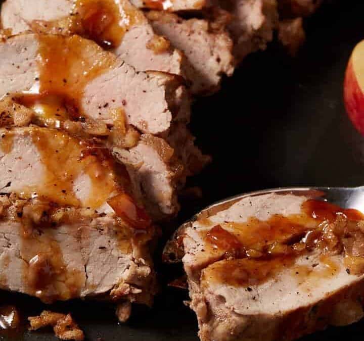 A platter with pork tenderloin and apples with a serving spoon.