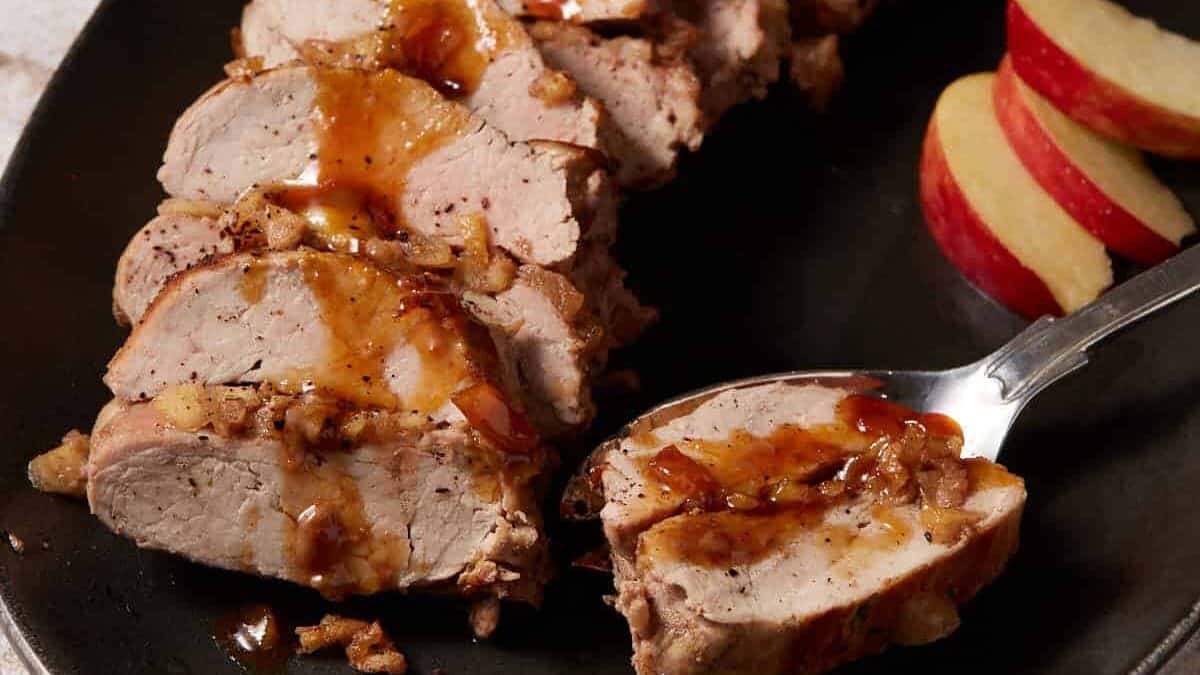 A platter with pork tenderloin and apples with a serving spoon.