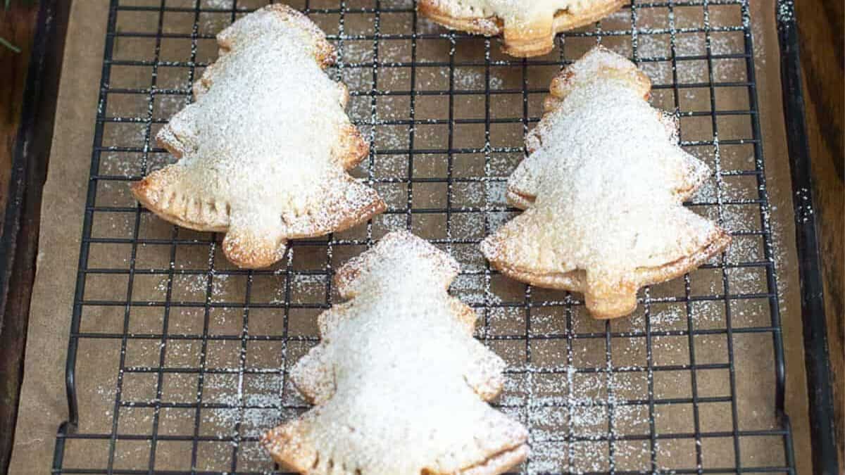 Five Christmas tree-shaped cookies, dusted with powdered sugar, are cooling on a wire rack placed over parchment paper. A copper Christmas tree-shaped cookie cutter is visible in the top-right corner. Evergreen branches can be seen in the background.